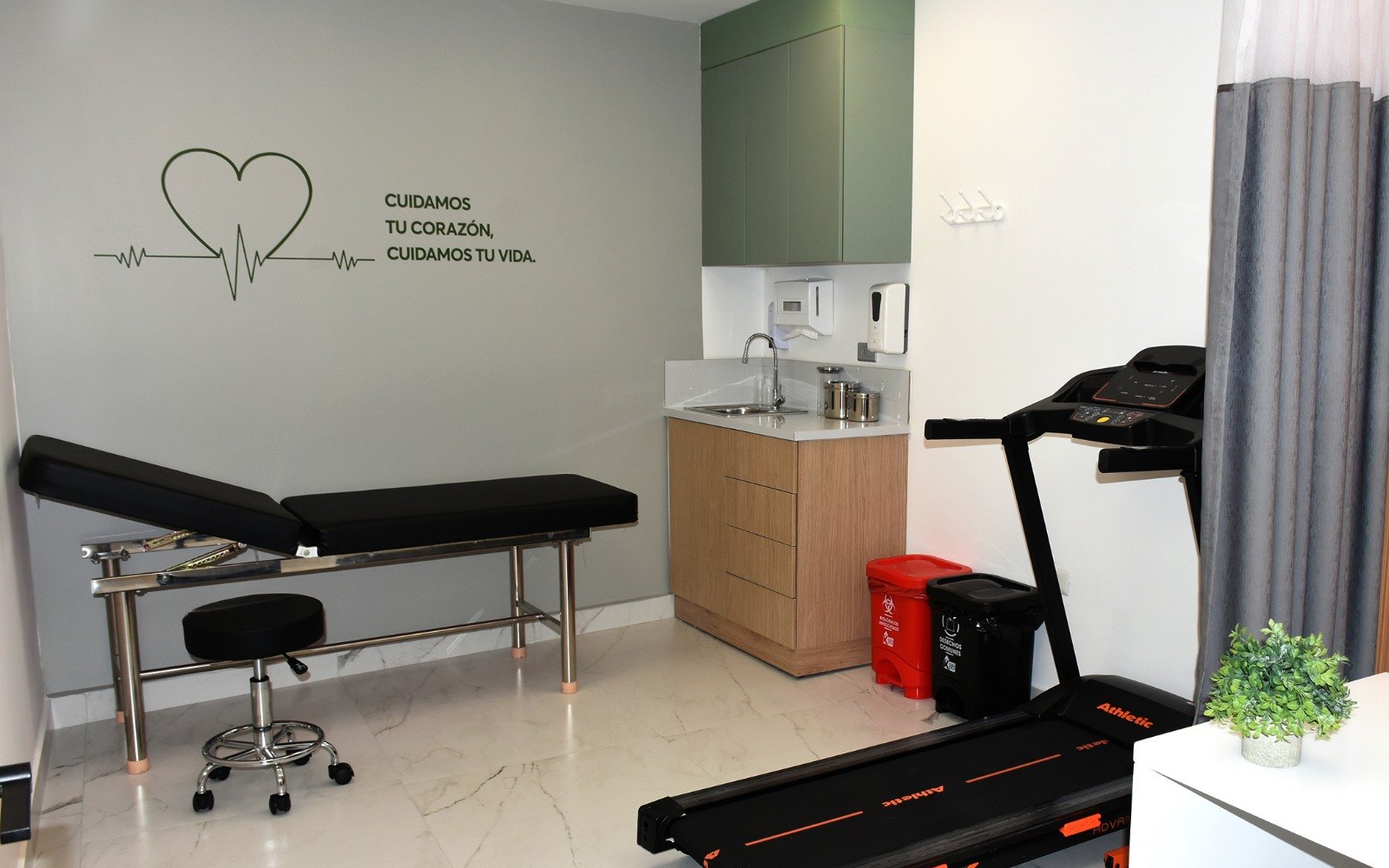 Consultorio de cardiología de CEMEDEC, con camilla, banda para prueba de esfuerzo y el mural «Cuidamos tu corazón, cuidamos tu vida»